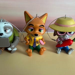 May include: Three Zootopia character ornaments: a sloth with a laptop, a fox with a tie and a pink donut, and a bunny with glasses and a carrot. Each ornament has a silver string for hanging.