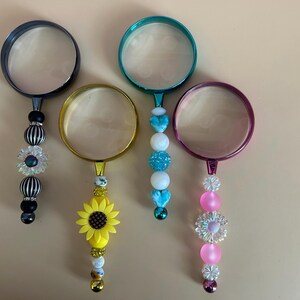 May include: Four decorative magnifying glasses with colorful beaded handles. The magnifying glasses have black, gold, teal, and purple frames. The handles feature a sunflower, hearts, and various beads. The lenses are clear.