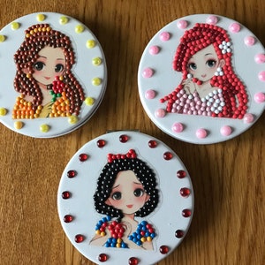 May include: Three round, white compact mirrors with images of Disney princesses. The first mirror features Belle with a yellow background, the second mirror features Ariel with a pink background, and the third mirror features Snow White with a red background.