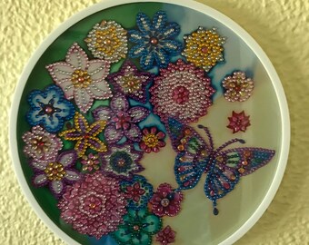 May include: A round, white-framed wall hanging with a colorful butterfly and floral design. The design is made of small, iridescent beads in shades of pink, purple, blue, green, and yellow.