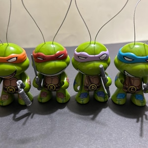 May include: Four green Teenage Mutant Ninja Turtle figurines with different colored masks and weapons. Each figure has a letter on its chest. They are ornaments with silver wire hangers.