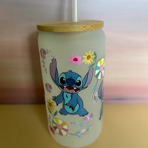 Op de afbeelding: Een matglazen beker met een blauw stripfiguur en bloemmotieven. Het heeft een bamboe deksel en een doorzichtig rietje. De beker is versierd met een stripfiguur en kleurrijke bloemen.