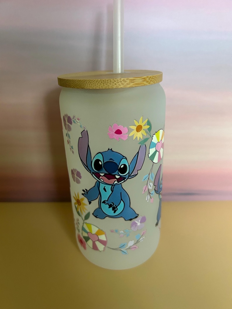 Op de afbeelding: Een matglazen beker met een blauw stripfiguur en bloemmotieven. Het heeft een bamboe deksel en een doorzichtig rietje. De beker is versierd met een stripfiguur en kleurrijke bloemen.