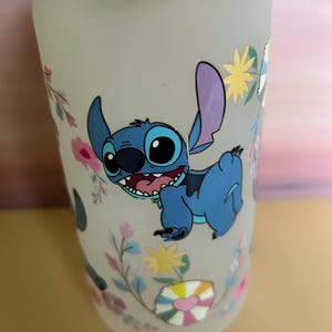 Op de afbeelding: Een matglazen beker met een blauw stripfiguur met grote ogen en een brede grijns. De beker is versierd met bloemen en geometrische ontwerpen in pastelkleuren en heeft een bamboe deksel.