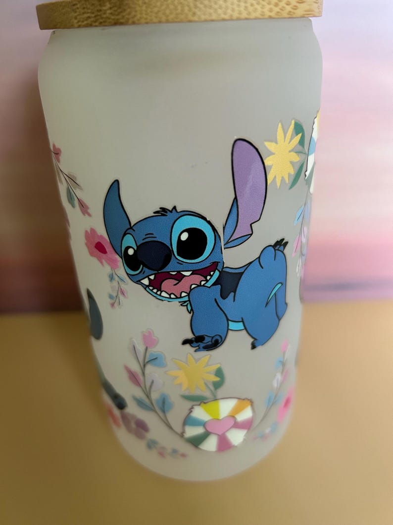 Op de afbeelding: Een matglazen beker met een blauw stripfiguur met grote ogen en een brede grijns. De beker is versierd met bloemen en geometrische ontwerpen in pastelkleuren en heeft een bamboe deksel.