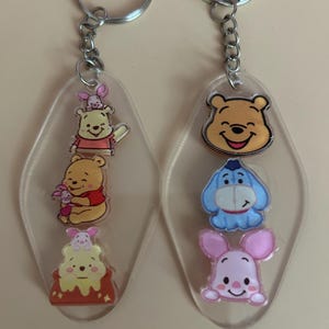 May include: Two clear acrylic keychains featuring cartoon characters. One keychain has Winnie the Pooh and Piglet, the other has Winnie the Pooh, Eeyore, and Piglet. Each keychain has a silver metal keyring and chain.