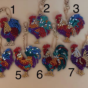 May include: Seven rooster-shaped keychains with colorful, sparkling embellishments. Each keychain features a different color scheme, including red, blue, green, and purple. The keychains have gold-colored hardware and are arranged on a light brown surface.