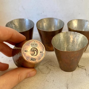 Può includere: Set di quattro bicchieri da shot in rame con finitura martellata. I bicchieri sono disposti a semicerchio con una moneta svedese da 5 öre del 1970 al centro.