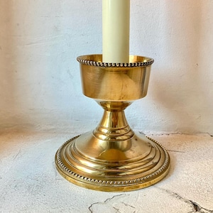 Può includere: Un portacandele dorato con un bordo perlato e una base rotonda. Una candela bianca è nel portacandele.