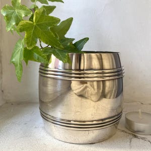 May include: A silver, cylindrical vase with three horizontal bands around the middle. The vase is sitting on a white surface with a small white candle in the background.