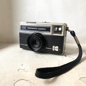 May include: A black and silver Kodak Instamatic camera with a black strap. The camera has a black lens and a white label that reads "77X Instamatic Camera".