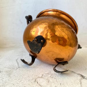 Puede incluir: Una olla de cobre martillado con cuerpo redondeado y borde acampanado. La olla tiene tres patas curvas y un asa negra. Un elemento decorativo negro est&aacute; unido al lateral. La olla probablemente se utiliza para cocinar o servir.