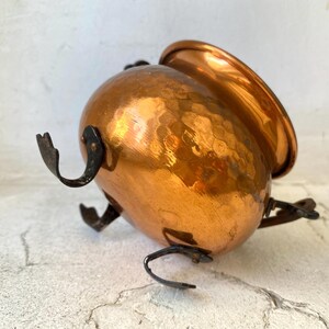 Puede incluir: Una olla de metal martillado de color cobre, con cuerpo redondeado y borde acampanado. La olla tiene tres patas curvas de metal oscuro y dos asas curvas. La superficie de la olla tiene un aspecto texturizado y con hoyuelos.