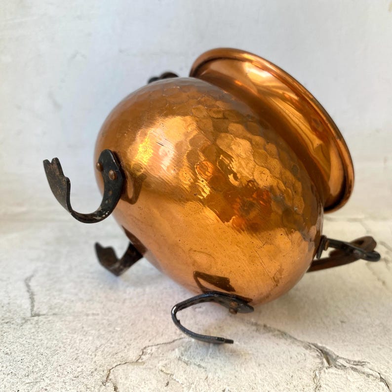 Puede incluir: Una olla de metal martillado de color cobre, con cuerpo redondeado y borde acampanado. La olla tiene tres patas curvas de metal oscuro y dos asas curvas. La superficie de la olla tiene un aspecto texturizado y con hoyuelos.