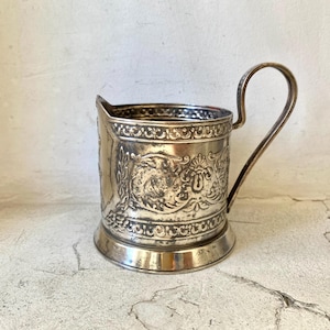 Könnte beinhalten: Silberfarbener Metall-Tassenhalter mit einem verzierten, geprägten Design. Der Tassenhalter hat einen Griff und ein dekoratives Muster um den Rand und den Sockel. Das Design umfasst florale und scrollartige Elemente, die ihm eine Vintage-Ästhetik verleihen.