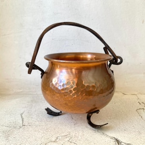 May include: A small, copper-colored cauldron with a hammered texture. It has a dark metal handle and three curved feet. The cauldron is designed with a rounded body and a wide opening, suitable for decorative or small-scale use.