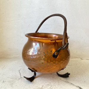 Puede incluir: Un peque&ntilde;o caldero de cobre martillado con un asa curva y tres patas negras. La olla de cobre tiene una superficie brillante y reflectante y una apariencia r&uacute;stica y vintage. El asa es de metal oscuro.