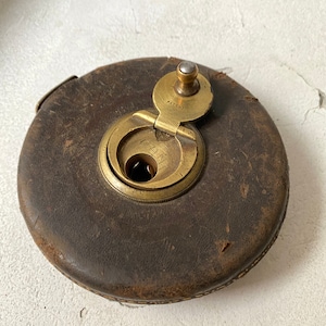 Puede incluir: Una cinta métrica vintage cubierta de cuero con un mecanismo de latón. La cinta métrica tiene una tapa con bisagras con la palabra "PATENT" inscrita en ella.