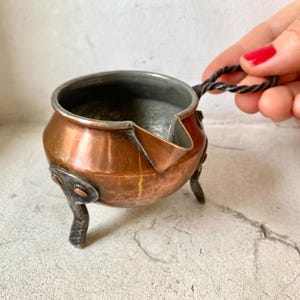 Könnte beinhalten: Ein kleiner Kupfertopf mit einem Henkel und drei Beinen. Der Topf hat eine gehämmerte Oberfläche und einen Ausguss. Der Topf steht auf einer weißen Oberfläche.