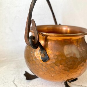 Puede incluir: Un peque&ntilde;o caldero de cobre martillado con un asa de metal oscuro y tres patas peque&ntilde;as. El caldero tiene una superficie brillante y reflectante y una apariencia r&uacute;stica y vintage. El asa est&aacute; sujeta con soportes de metal oscuro.