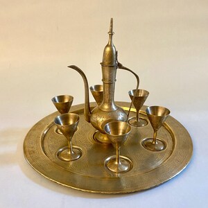 May include: A vintage brass tea set, including a tall, ornate pitcher with a slender spout and six small goblets, all resting on a round, engraved tray. The set has a warm, golden hue and intricate floral patterns.
