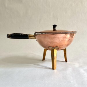 Könnte beinhalten: Eine Kupfer-Chafing Dish mit einem schwarzen Griff und einem Messingständer. Die Schale hat einen Deckel mit einem schwarzen Knopf.