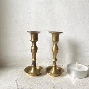 Peut inclure: Deux bougeoirs anciens en laiton de conception classique. Les bougeoirs ont une base ronde, une tige élancée et un sommet évasé. Une bougie chauffe-plat blanche est à droite. Les bougeoirs sont sur une surface blanche.