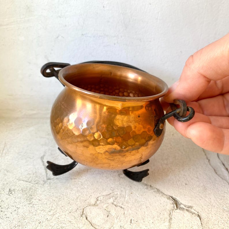Puede incluir: Un peque&ntilde;o caldero de cobre martillado con cuerpo redondo y dos asas en forma de lazo. El caldero se apoya sobre un soporte de metal negro con tres patas. El cobre tiene un brillo c&aacute;lido y reflectante.