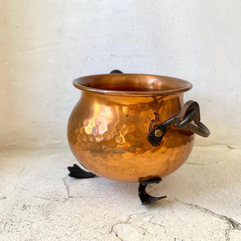 Puede incluir: Un peque&ntilde;o caldero de cobre martillado con un cuerpo redondeado y tres patas negras. El caldero tiene dos asas negras y una superficie brillante y reflectante. El interior es oscuro y el exterior tiene un aspecto texturizado y con hoyuelos.