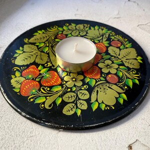 Peut inclure: Un bougeoir en métal noir avec un motif floral composé de fraises rouges, de feuilles vertes et d'accents dorés. Une bougie chauffe-plat blanche est placée au centre du bougeoir.
