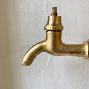 Może przedstawiać: Zabytkowy kran z mosiądzu z zakrzywioną wylewką i kwadratowym uchwytem. Kran ma wygląd postarzałego, ze śladami wieku i użytkowania. Mosiądz ma ciepły, złoty odcień, a ogólny design jest prosty i funkcjonalny.