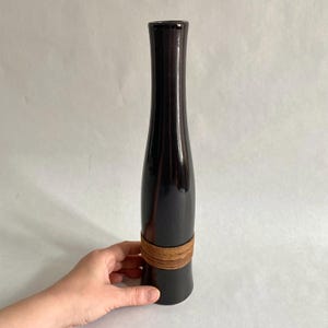 Puede incluir: Un jarrón de cerámica negro con una banda tejida marrón alrededor del medio. El jarrón tiene un cuello estrecho y una base ancha.