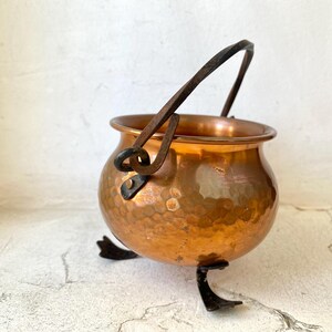 Puede incluir: Un peque&ntilde;o caldero de cobre martillado con un asa de metal oscuro y tres patas peque&ntilde;as. El caldero tiene un cuerpo redondeado y una superficie brillante y reflectante. El asa es arqueada y las patas son curvas.