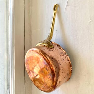 Könnte beinhalten: Ein Kupfertopf mit Messinggriff und dekorativen Details. Der Topf hat einen abgerundeten Boden und hängt an einer weißen Wand. Der Griff ist mit verzierten Messingdetails befestigt.