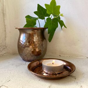 Könnte beinhalten: Eine gehämmerte Kupfervase mit einem kleinen, runden, gehämmerten Kupfer-Kerzenhalter und einem weißen Teelicht. Die Vase ist mit grünem Efeu gefüllt.