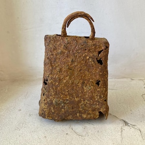 Könnte beinhalten: Eine rechteckige, rostige Metallglocke mit gebogenem Griff. Die Glocke hat eine verwitterte, strukturierte Oberfläche mit mehreren kleinen Löchern. Die Gesamtfarbe ist eine Mischung aus Braun und Orange, was auf Alter und Witterungseinfluss hindeutet.