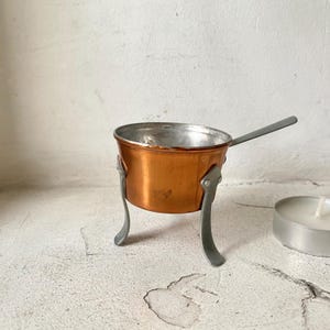 Può includere: Una piccola pentola di rame con un manico e tre gambe. La pentola è su una superficie bianca.