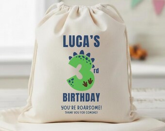 May include: A natural canvas drawstring bag with the text "LUCA'S 3rd BIRTHDAY" in blue and green. The number 3 is designed as a green dinosaur. Below the text is "YOU'RE ROARSOME! THANK YOU FOR COMING!" The bag is sitting on a folded canvas pouch.