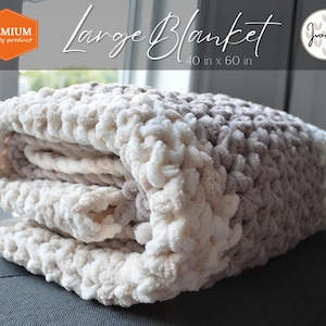May include: A large, crocheted blanket in shades of beige and brown. The blanket is folded and displayed on a gray couch. The blanket is 40 inches by 60 inches.