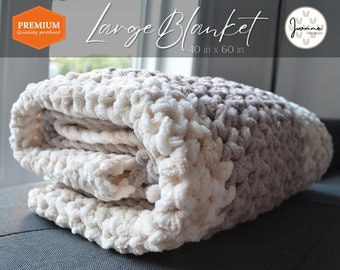 May include: A large, crocheted blanket in shades of beige and brown. The blanket is folded and displayed on a gray couch. The blanket is 40 inches by 60 inches.