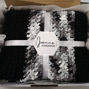May include: A black, gray, and white striped crocheted blanket is folded and tied with a white ribbon in a white gift box. A white tag with the text "Joanne Handmade" is attached to the ribbon.