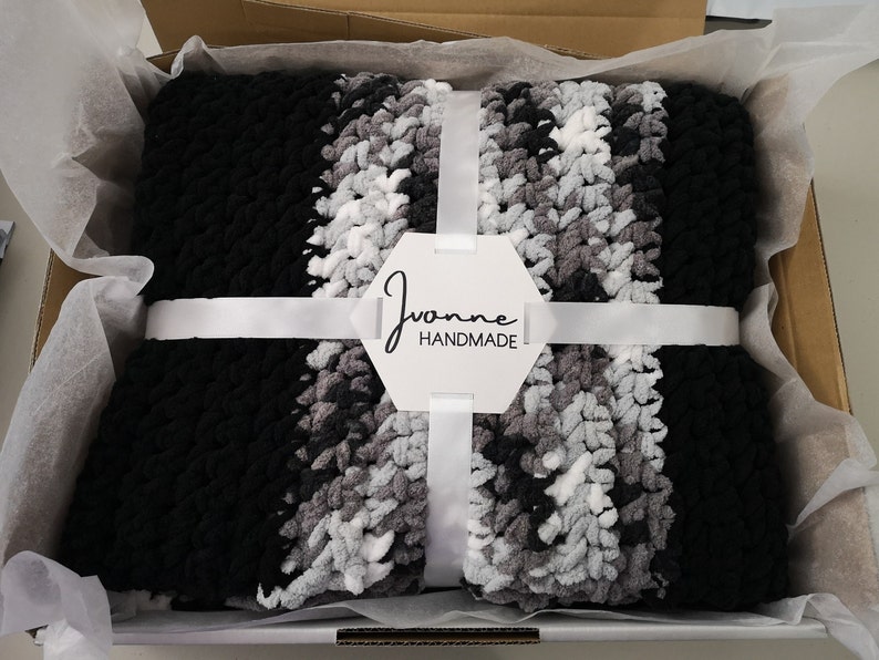 May include: A black, gray, and white striped crocheted blanket is folded and tied with a white ribbon in a white gift box. A white tag with the text "Joanne Handmade" is attached to the ribbon.