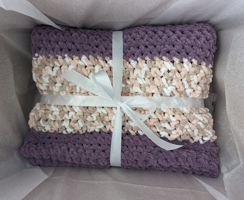 K&ouml;nnte beinhalten: Eine gestrickte Decke in Lila und Rosa mit einem wei&szlig;en Band darum gebunden.