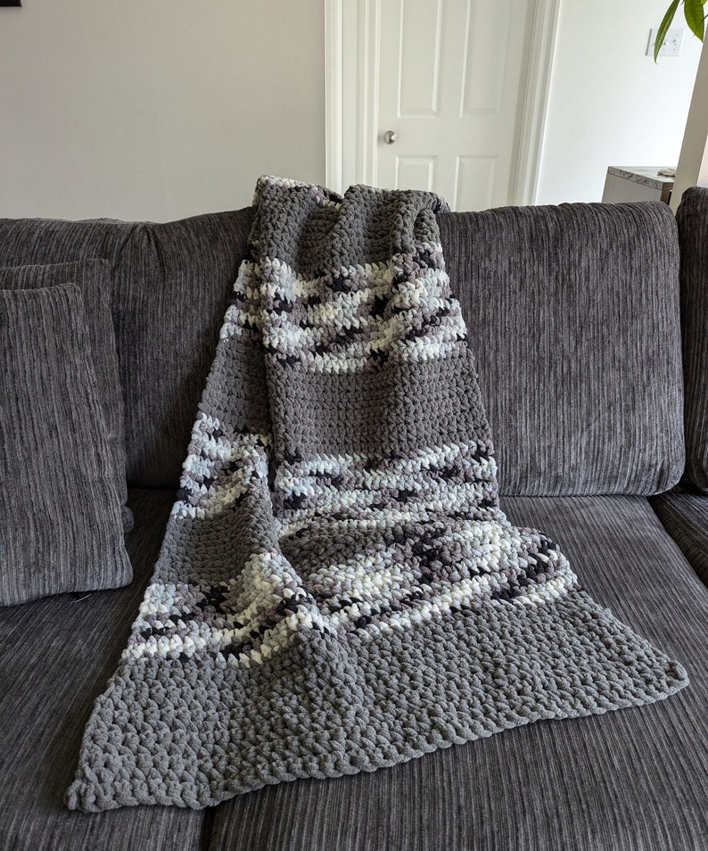 K&ouml;nnte beinhalten: Eine graue und wei&szlig;e gestreifte geh&auml;kelte Wolldecke mit einer strukturierten, flauschigen Optik. Die Decke liegt &uuml;ber einem grauen Sofa.