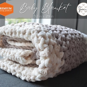 Puede incluir: Una suave manta de bebé de crochet en color crema y marrón, doblada en un sofá gris. La manta mide 81 cm por 74 cm. El texto "Baby Blanket" y "32 in x 29 in" está impreso sobre un fondo blanco.