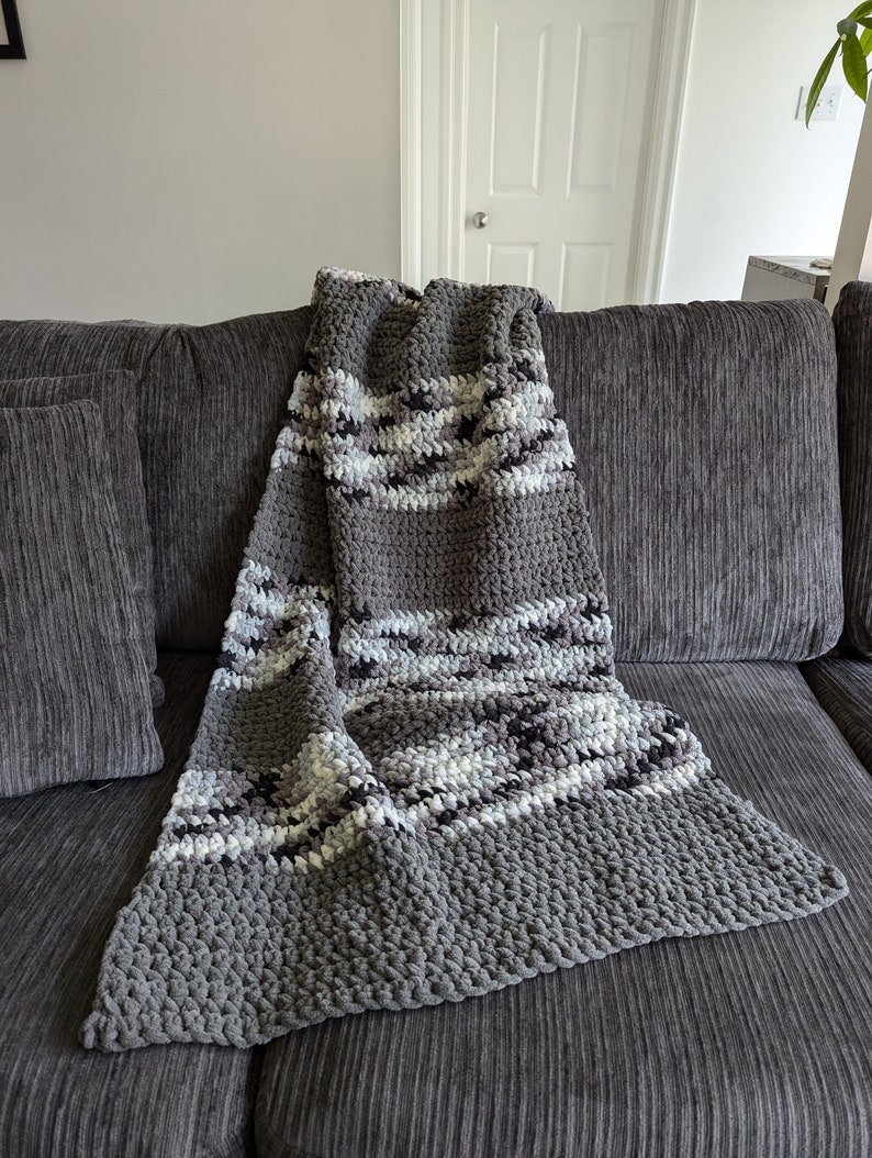May include: A gray and white striped crocheted throw blanket with a textured, fluffy surface. The blanket is draped over a gray couch.