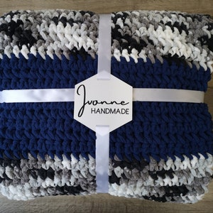 May include: A navy blue, gray, black, and white striped crocheted blanket with a white ribbon tied around it. The blanket has a white tag with the text "Joanne Handmade" on it.