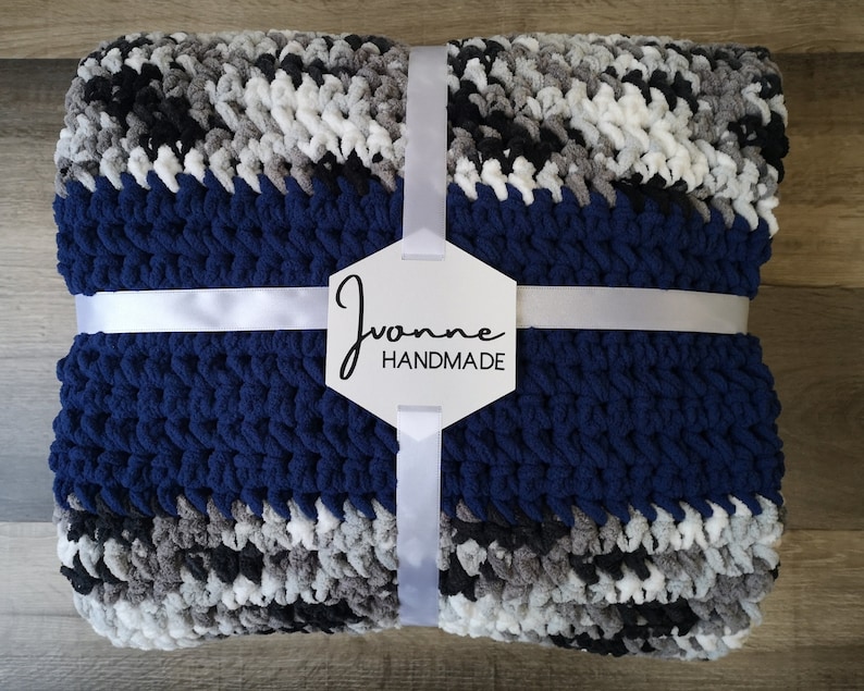 May include: A navy blue, gray, black, and white striped crocheted blanket with a white ribbon tied around it. The blanket has a white tag with the text "Joanne Handmade" on it.