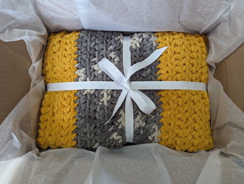 K&ouml;nnte beinhalten: Eine geh&auml;kelte Decke in Gelb und Grau, gebunden mit einem wei&szlig;en Band, verpackt in einer mit wei&szlig;em Seidenpapier ausgekleideten Schachtel.