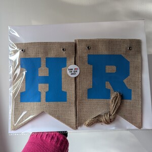 Puede incluir: Una pancarta de arpillera con las letras "HR" en azul sobre un fondo beige. La pancarta est&aacute; unida con un cordel y tiene una peque&ntilde;a etiqueta que dice "&iexcl;Gracias por tu pedido!" con un coraz&oacute;n rojo.